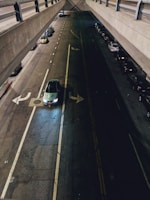 A dimly lit urban street with a view from above, showing a car with headlights on navigating an intersection beneath a concrete overpass. There are parked cars lining the street on both sides, with some visible in the shadows. The road appears well-marked, with lane lines and directional arrows indicating traffic flow.