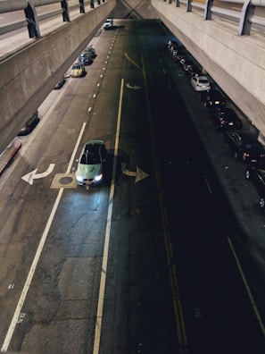 A dimly lit urban street with a view from above, showing a car with headlights on navigating an intersection beneath a concrete overpass. There are parked cars lining the street on both sides, with some visible in the shadows. The road appears well-marked, with lane lines and directional arrows indicating traffic flow.