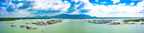A wide expanse of water with floating structures, possibly fishing farms or houses. There are lush green hills in the background under a partly cloudy sky, creating a serene and expansive landscape.