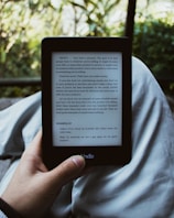 A person holds an e-reader device displaying a page of text. The background shows a blurred view of outdoor greenery with sunlight filtering through leaves. The person is seated, with the device resting on their lap.