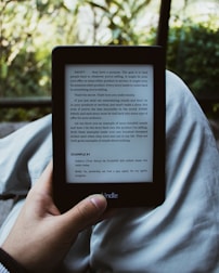 A person holds an e-reader device displaying a page of text. The background shows a blurred view of outdoor greenery with sunlight filtering through leaves. The person is seated, with the device resting on their lap.