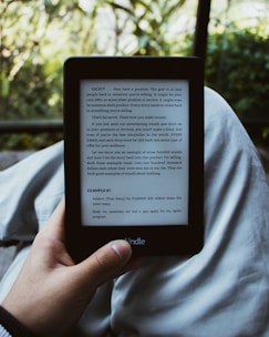 A person holds an e-reader device displaying a page of text. The background shows a blurred view of outdoor greenery with sunlight filtering through leaves. The person is seated, with the device resting on their lap.