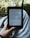 A person holds an e-reader device displaying a page of text. The background shows a blurred view of outdoor greenery with sunlight filtering through leaves. The person is seated, with the device resting on their lap.