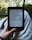 A person holds an e-reader device displaying a page of text. The background shows a blurred view of outdoor greenery with sunlight filtering through leaves. The person is seated, with the device resting on their lap.