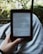 A person holds an e-reader device displaying a page of text. The background shows a blurred view of outdoor greenery with sunlight filtering through leaves. The person is seated, with the device resting on their lap.