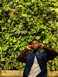 A person is standing in front of a lush green vertical garden wall, wearing headphones with eyes closed, suggesting they are enjoying music. The wall is dense with a variety of leaves and plants, creating a vibrant natural backdrop.