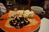 A dessert plate featuring a crepe with chocolate sauce and almond slivers, accompanied by two scoops of vanilla ice cream topped with whipped cream, is presented. A spoon rests on the plate, and in the background, there is a cup of tea and a glass of water on a wooden table.