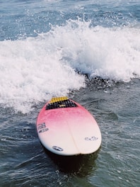 A surfboard with a pink to white gradient sits on the water amidst splashing waves. The board features various decals and a traction pad near the tail. The surrounding water shows a mix of calm areas and energetic white foam from the waves.