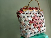 A woven handbag made from recycled materials, featuring a grid pattern with red, white, and beige elements. The bag's structure is reinforced by woven handles, creating a rustic and eco-friendly appearance.