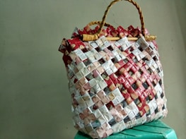 A woven handbag made from recycled materials, featuring a grid pattern with red, white, and beige elements. The bag's structure is reinforced by woven handles, creating a rustic and eco-friendly appearance.