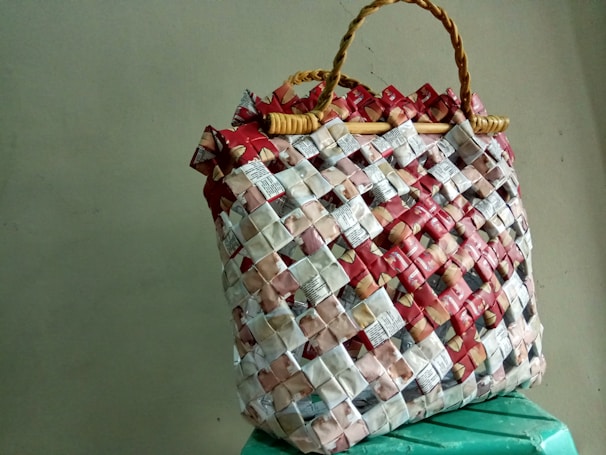 A woven handbag made from recycled materials, featuring a grid pattern with red, white, and beige elements. The bag's structure is reinforced by woven handles, creating a rustic and eco-friendly appearance.