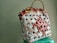 A woven handbag made from recycled materials, featuring a grid pattern with red, white, and beige elements. The bag's structure is reinforced by woven handles, creating a rustic and eco-friendly appearance.