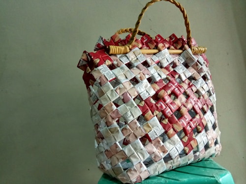 A woven handbag made from recycled materials, featuring a grid pattern with red, white, and beige elements. The bag's structure is reinforced by woven handles, creating a rustic and eco-friendly appearance.