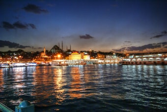 The image captures a scenic waterfront view during dusk or early evening, with a city skyline and a prominent mosque in the background. The calm water reflects the vibrant lights from the shoreline, where several buildings and boats can be seen. The sky features a gradient from deep blue to soft orange as the sun sets.