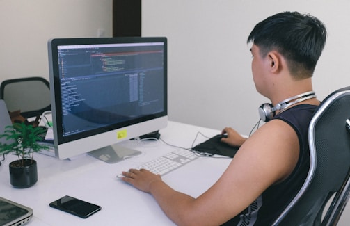 A person is sitting at a desk using a computer with code displayed on the monitor. The individual is wearing headphones around their neck and appears focused on the task. The workspace includes a keyboard, mouse, smartphone, and a small potted plant.