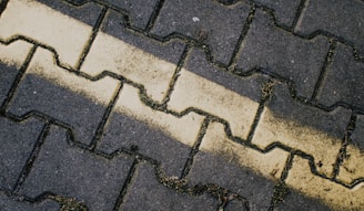 A close-up view of interlocking pavement bricks organized in a zigzag pattern. A faded yellow line runs diagonally across the bricks, with some patches of grass and dirt in the crevices.