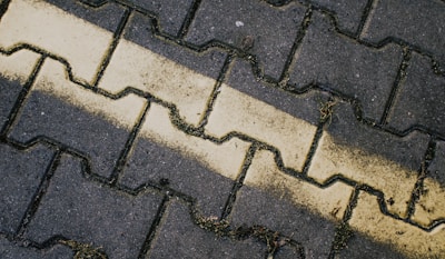 A close-up view of interlocking pavement bricks organized in a zigzag pattern. A faded yellow line runs diagonally across the bricks, with some patches of grass and dirt in the crevices.