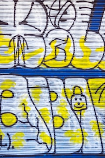 Graffiti art on a metal shutter featuring abstract shapes and figures in black outlines with bright yellow highlights against a white background. Some parts include thick lines and circular designs.
