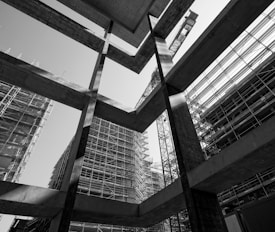 A black and white photograph capturing the geometric lines and forms of construction in progress. The image features scaffolding surrounding partially built structures, with a large crane in the background. The high contrast highlights the textures and materials used in modern urban architecture.