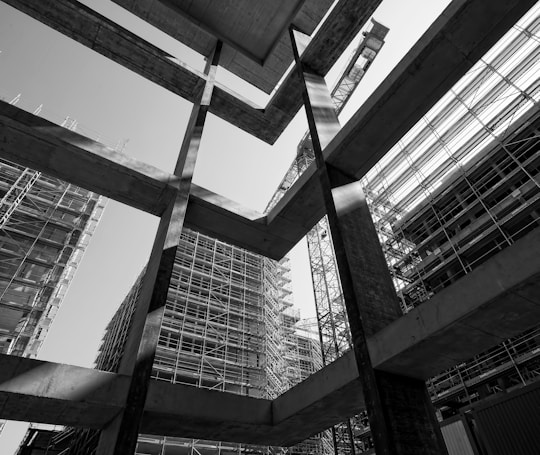 A black and white photograph capturing the geometric lines and forms of construction in progress. The image features scaffolding surrounding partially built structures, with a large crane in the background. The high contrast highlights the textures and materials used in modern urban architecture.