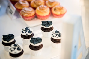 A display of baked goods featuring six chocolate cupcakes, each decorated with black and white character designs on top. Behind the cupcakes, a tray of golden-brown pastries, likely cinnamon rolls, is visible. The setup suggests a bakery or a dessert-themed event.