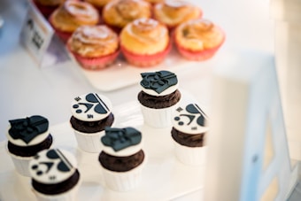 A display of baked goods featuring six chocolate cupcakes, each decorated with black and white character designs on top. Behind the cupcakes, a tray of golden-brown pastries, likely cinnamon rolls, is visible. The setup suggests a bakery or a dessert-themed event.
