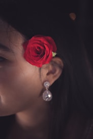 A close-up of a person's profile focuses on an ear adorned with a large gem-encrusted earring and a vibrant red rose placed above the ear. The background is dark, emphasizing the bright red of the rose and the sparkling gem of the earring.