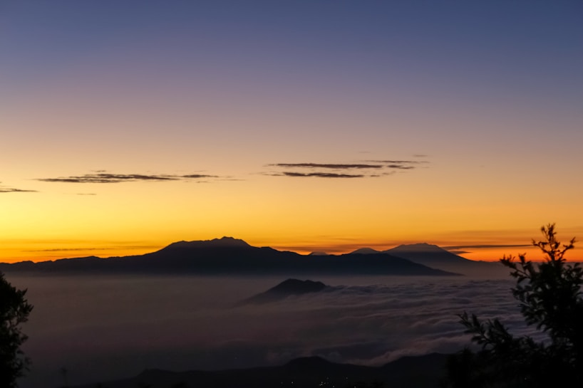 The image features a stunning sunrise or sunset over a mountainous landscape. The sky transitions beautifully from a deep blue at the top to a warm golden yellow near the horizon, with a few scattered clouds adding texture. The silhouette of the mountains is prominently visible against the colorful sky, and a layer of mist or clouds hugs the valleys between the peaks.