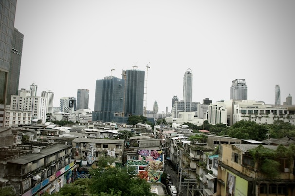 A cityscape featuring a mix of modern skyscrapers and older, densely packed buildings in the foreground. Several tall buildings under construction are visible, surrounded by cranes. Graffiti covers the lower level buildings, adding an urban, gritty atmosphere.