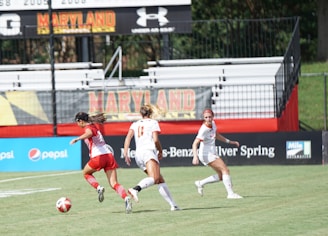 Three female soccer players are active on a soccer field. One player in a red uniform is dribbling a soccer ball, while two players in white uniforms are running towards her. The game appears to be taking place in a stadium with mostly empty stands and banners promoting Maryland and various sponsors around the field.