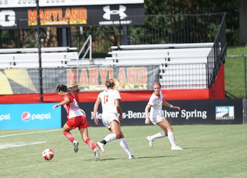 Three female soccer players are active on a soccer field. One player in a red uniform is dribbling a soccer ball, while two players in white uniforms are running towards her. The game appears to be taking place in a stadium with mostly empty stands and banners promoting Maryland and various sponsors around the field.