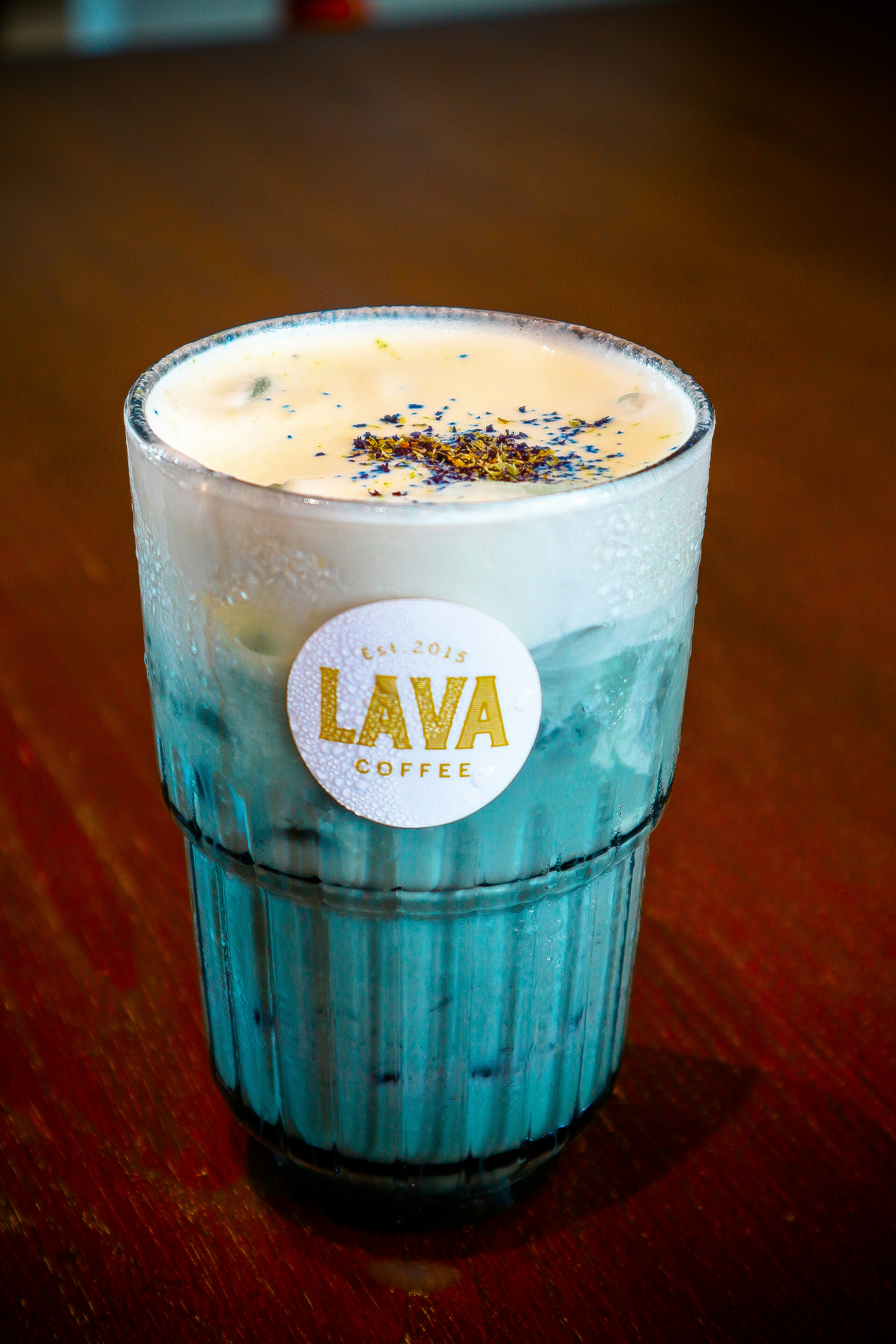 A creamy ube latte served in a clear glass, topped with a swirl of foam and a sprinkle of purple powder.