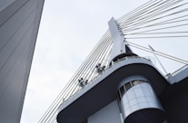A modern architectural structure featuring a bridge or tower with steel cables extending from a central pillar. The design includes circular metal fixtures and an angular perspective that highlights the sleek lines and geometric shapes.