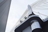 A modern architectural structure featuring a bridge or tower with steel cables extending from a central pillar. The design includes circular metal fixtures and an angular perspective that highlights the sleek lines and geometric shapes.