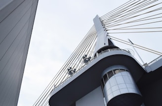 A modern architectural structure featuring a bridge or tower with steel cables extending from a central pillar. The design includes circular metal fixtures and an angular perspective that highlights the sleek lines and geometric shapes.