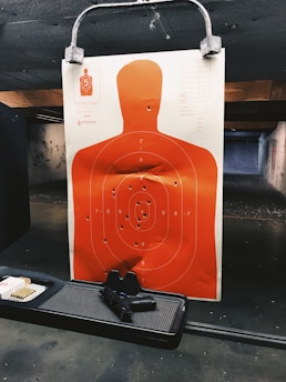 A shooting range target features a human silhouette with several bullet holes. The target is a bright orange color and is secured to a holder. Below the target, there is a black handgun placed on a tray next to a box of ammunition. The shooting range has a dim, industrial atmosphere with concrete walls and a paneled ceiling.