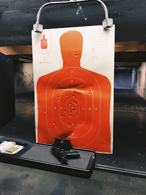 A shooting range target features a human silhouette with several bullet holes. The target is a bright orange color and is secured to a holder. Below the target, there is a black handgun placed on a tray next to a box of ammunition. The shooting range has a dim, industrial atmosphere with concrete walls and a paneled ceiling.