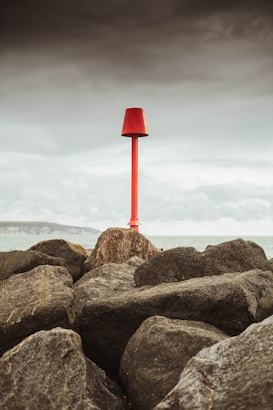 A red navigation marker stands prominently atop a cluster of large, dark rocks. The backdrop features a calm sea and an overcast sky, creating a contrast between the marker and the muted tones of the environment.