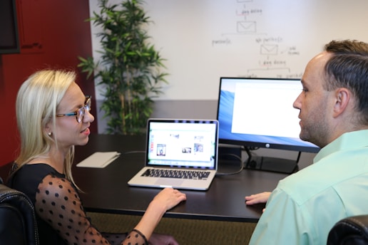 Two people are sitting at a desk engaged in conversation. A woman with long blonde hair and glasses is facing a man with short hair, both appear to be in a professional setting. A laptop and a monitor displaying content related to their discussion are on the desk. A plant is positioned in the corner of the office providing a touch of greenery.