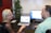 Two people are sitting at a desk engaged in conversation. A woman with long blonde hair and glasses is facing a man with short hair, both appear to be in a professional setting. A laptop and a monitor displaying content related to their discussion are on the desk. A plant is positioned in the corner of the office providing a touch of greenery.