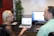 Two people are sitting at a desk engaged in conversation. A woman with long blonde hair and glasses is facing a man with short hair, both appear to be in a professional setting. A laptop and a monitor displaying content related to their discussion are on the desk. A plant is positioned in the corner of the office providing a touch of greenery.