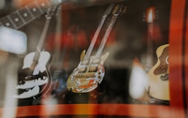Multiple guitars are displayed behind glass in a shop, with reflections creating a layered effect. The instruments vary in design, highlighting their unique styles and craftsmanship.