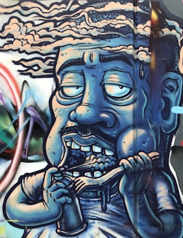 A graffiti-style artwork depicting a caricature of a human figure with a blue hue. The figure has exaggerated facial features, with half-closed, large eyes, a mustache, and showing teeth while holding a spoon and a cup. The background includes abstract shapes and vibrant colors, giving a dynamic feel to the mural.