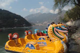 Colorful dragon-shaped paddle boats are lined up along the edge of a calm lake. Surrounding the water are lush green hills, topped with a few scattered clouds against a blue sky. The scene has a peaceful and inviting atmosphere.