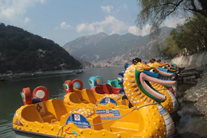 Colorful dragon-shaped paddle boats are lined up along the edge of a calm lake. Surrounding the water are lush green hills, topped with a few scattered clouds against a blue sky. The scene has a peaceful and inviting atmosphere.