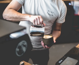 A person wearing a t-shirt is skillfully pouring milk from a metal pitcher into a cup of coffee, creating latte art. The setting appears to be a coffee shop with visible equipment and a relaxed ambiance.