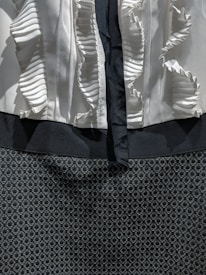 A close-up view of a garment with a ruffled white fabric on the top section and a dark, textured, and patterned fabric on the lower section. The ruffles create a layered effect while the dark fabric has a circular geometric design.