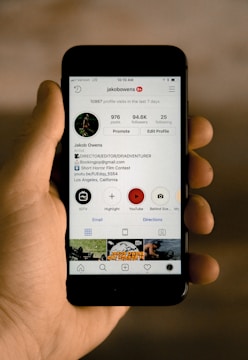 A hand is holding a smartphone displaying an Instagram profile. The profile belongs to a user named jakobowens, featuring a circular profile picture, user stats, and several icons for various Instagram functions. The screen shows text denoting the number of posts, followers, and following, along with buttons for promoting and editing the profile. Below the profile information are icons for IGTV, highlights, and YouTube, among others. The background is blurred, focusing attention on the phone and its screen.