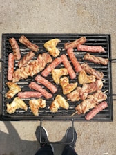 Various types of meat, including sausages, chicken wings, and skewered pieces, are being grilled on a black barbecue grill. The grill is placed on a textured ground surface and there's a person standing in front of it, visible only from the waist down and wearing dark shoes.