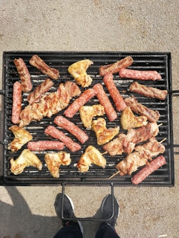 Various types of meat, including sausages, chicken wings, and skewered pieces, are being grilled on a black barbecue grill. The grill is placed on a textured ground surface and there's a person standing in front of it, visible only from the waist down and wearing dark shoes.